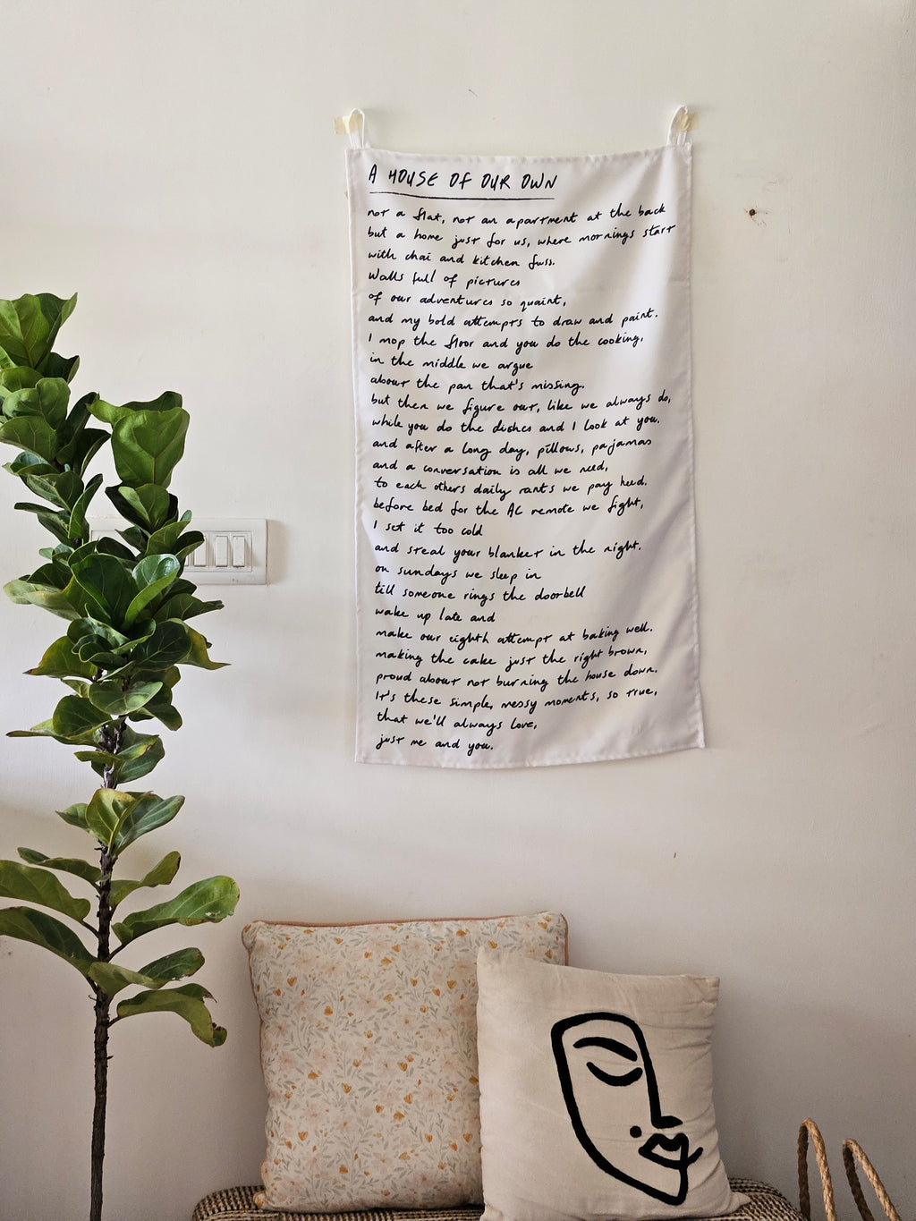 Cozy cotton wall art featuring a house and text embroidery, styled above a wooden console with plants and cushions

