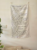 Minimalist beige cotton wall hanging with embroidered house illustration and quote, displayed on a clean white background.

